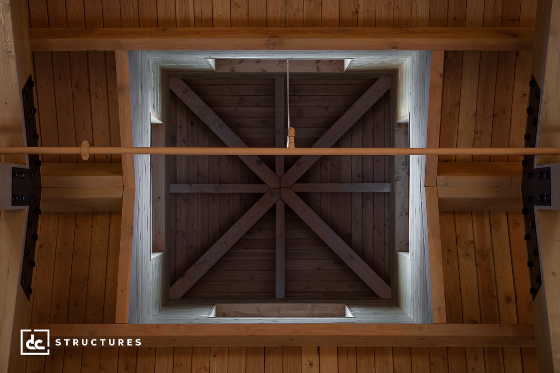 A geometric view looking up at a wooden ceiling with exposed beams in a square pattern and central cross-brace. Light enters through gaps.