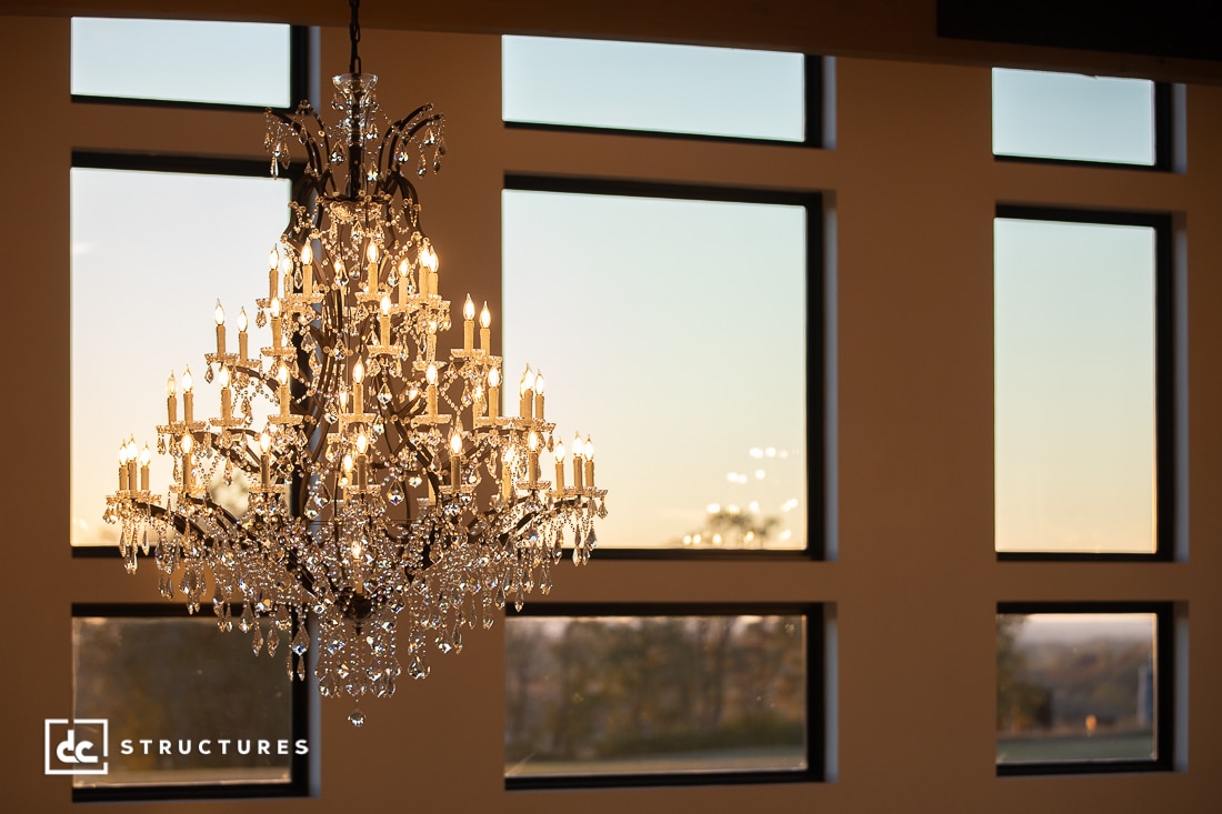 A large, ornate crystal chandelier glows with numerous lights in front of tall windows at sunset.