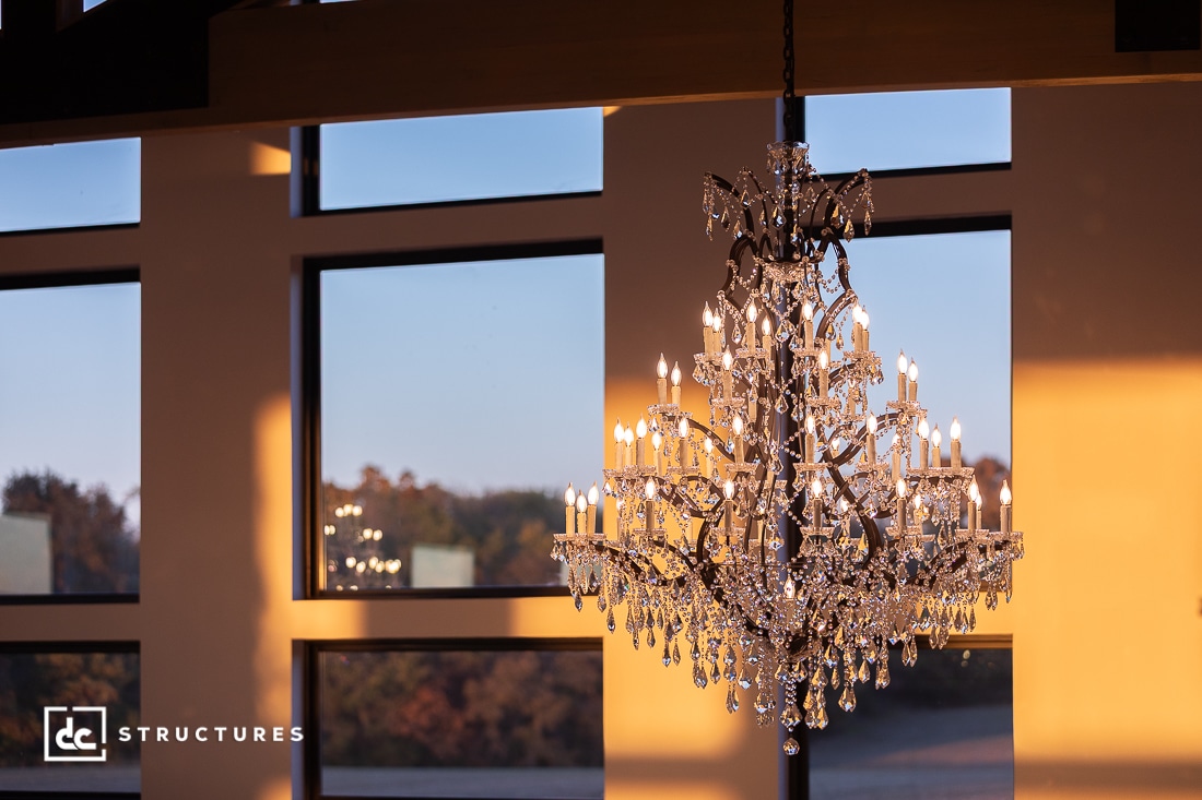 A large, ornate crystal chandelier hangs in front of tall windows, illuminated by natural light. Trees and the sunset are visible outside.