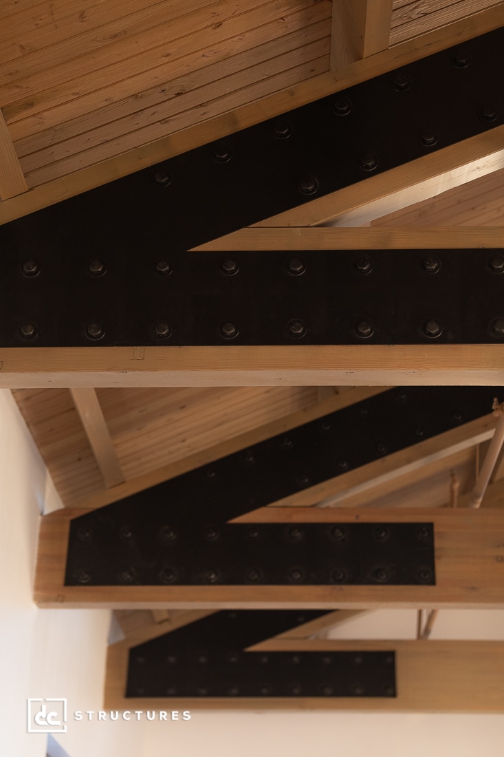 Exposed wooden ceiling beams with black metal plates and bolts form a zigzag pattern, showcasing an industrial architectural design.