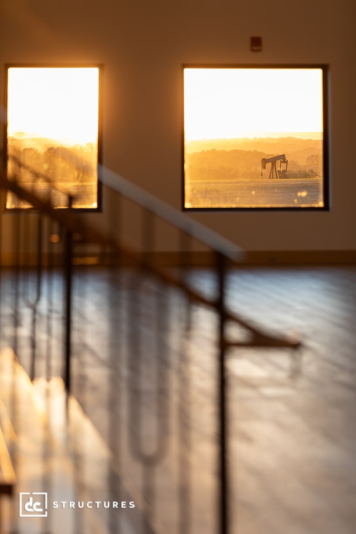 Sunlight streams through large windows, lighting an empty room with wooden floors. An oil pumpjack sits in a golden field outside.