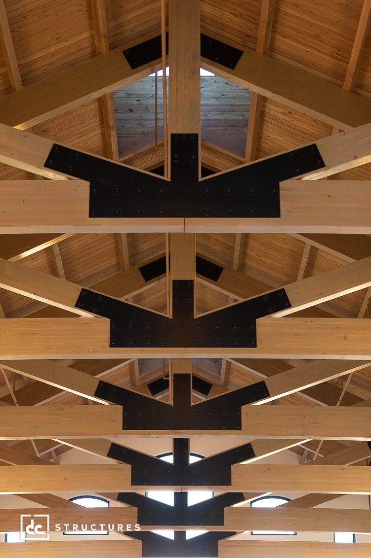 The image shows the interior ceiling of a building with exposed wooden beams, black metal fasteners, warm wood finish, and skylights above.