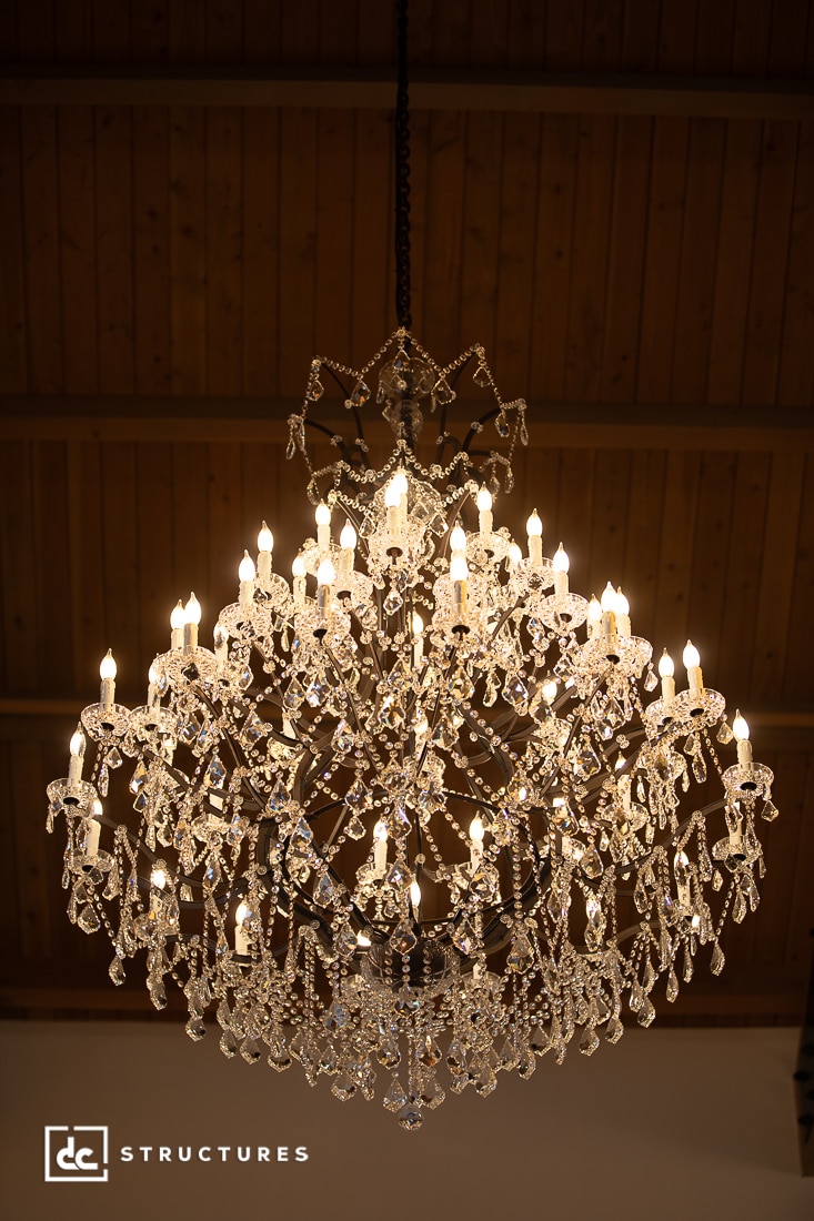 A large, ornate crystal chandelier with numerous lit bulbs hangs from a wooden ceiling, sparkling with luxurious elegance.