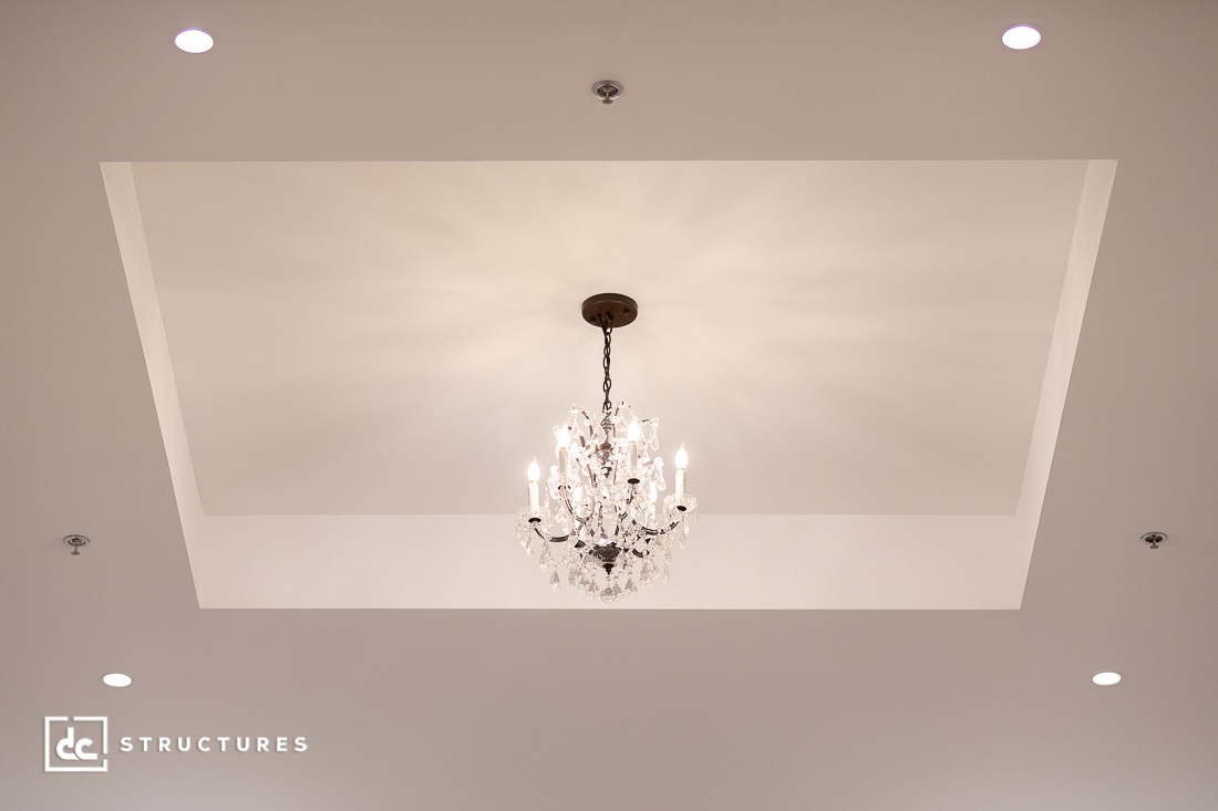 A crystal chandelier hangs from the center of a recessed white ceiling, with soft light radiating outward. Four recessed lights are installed at the corners.