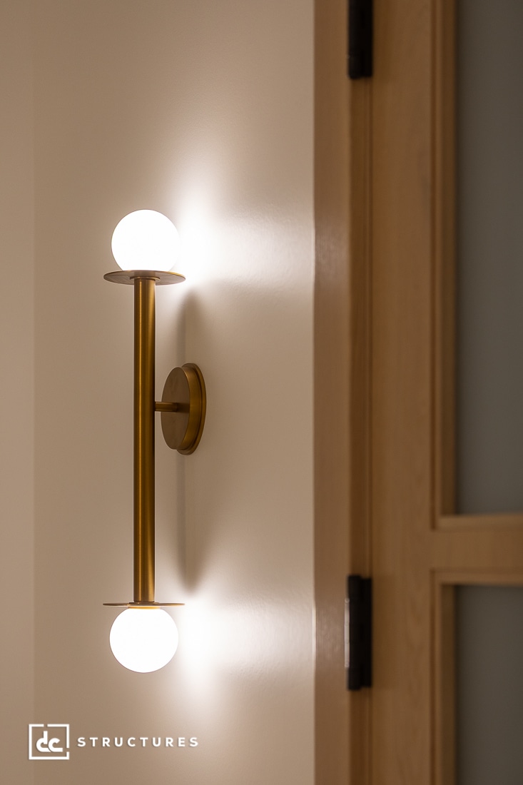 A modern brass wall sconce with two round white bulbs, mounted vertically on a light-colored wall next to a wooden door. The fixture emits a soft, warm light.