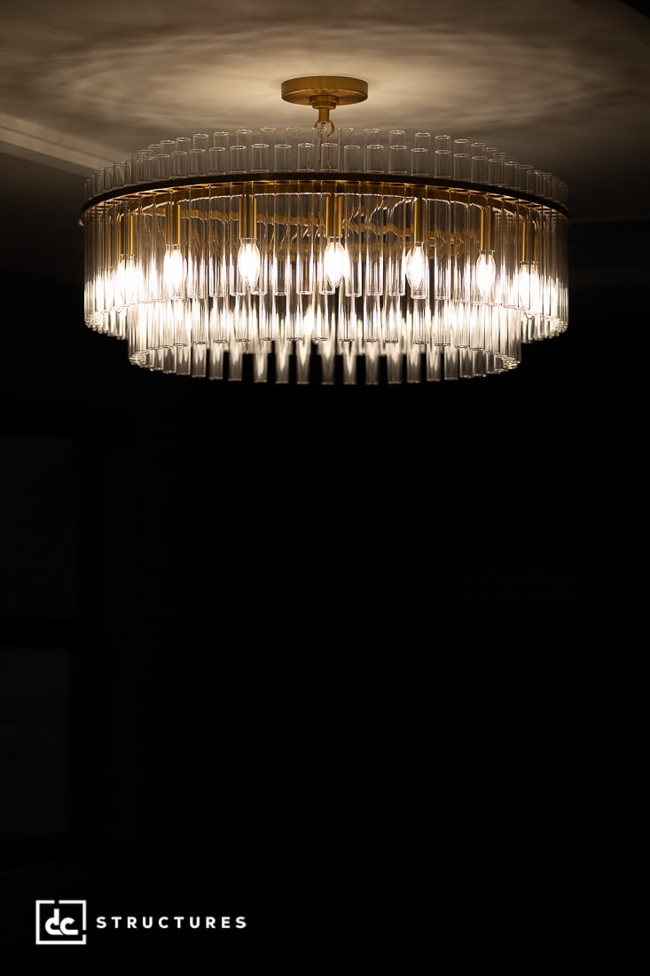 A modern chandelier with multiple exposed bulbs and glass rods hangs from the ceiling, casting a warm light. The fixture has a gold frame and is set against a dark background.