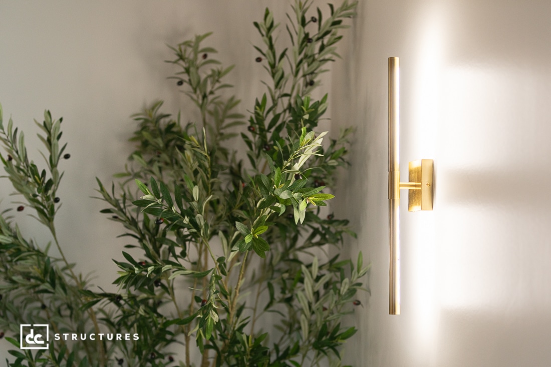 A modern gold wall sconce emits soft light on a white wall beside a leafy green plant.