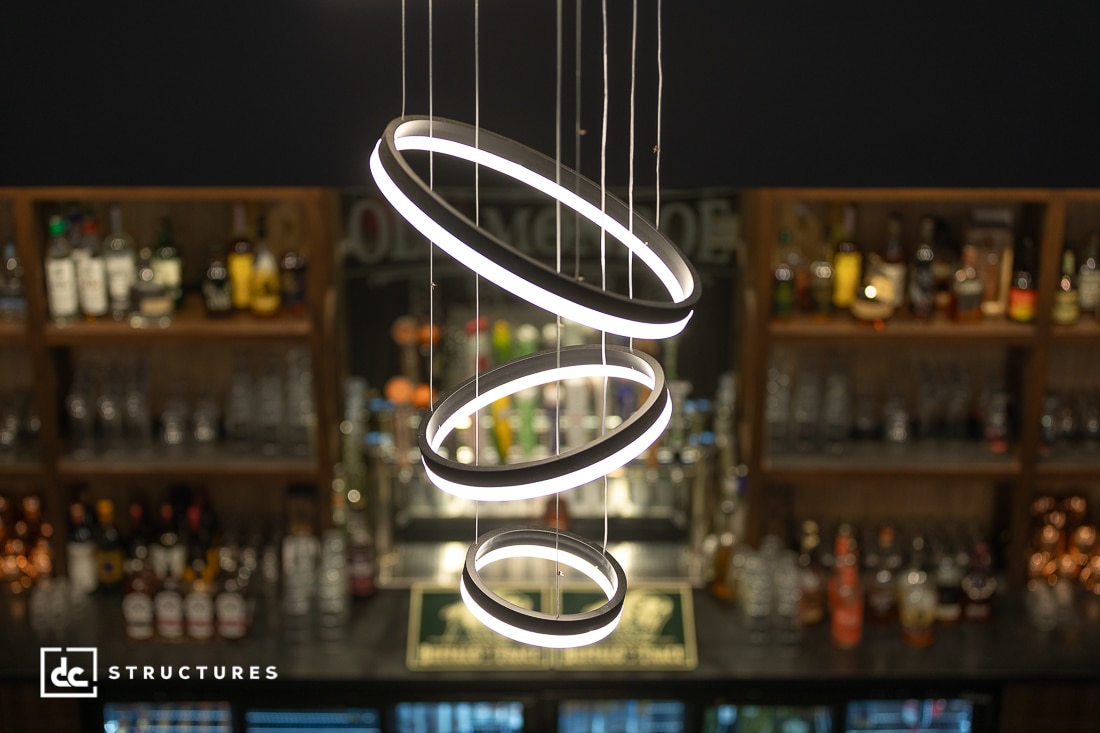 Modern circular pendant lights hang above a bar with shelves filled with bottles and glassware. The lights are glowing, creating a stylish and inviting atmosphere.