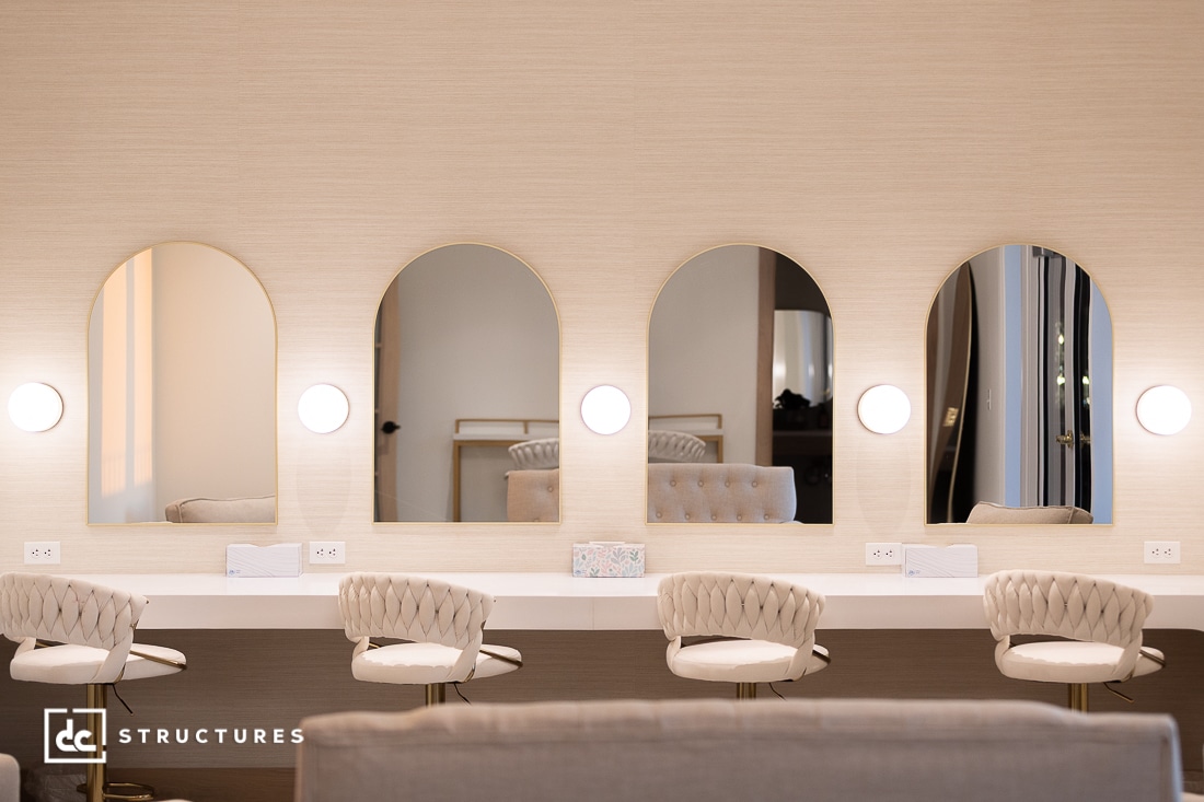 A modern vanity area with four tufted chairs at a long counter, each with a round wall-mounted light and an arched mirror. Decor is in soft beige tones.