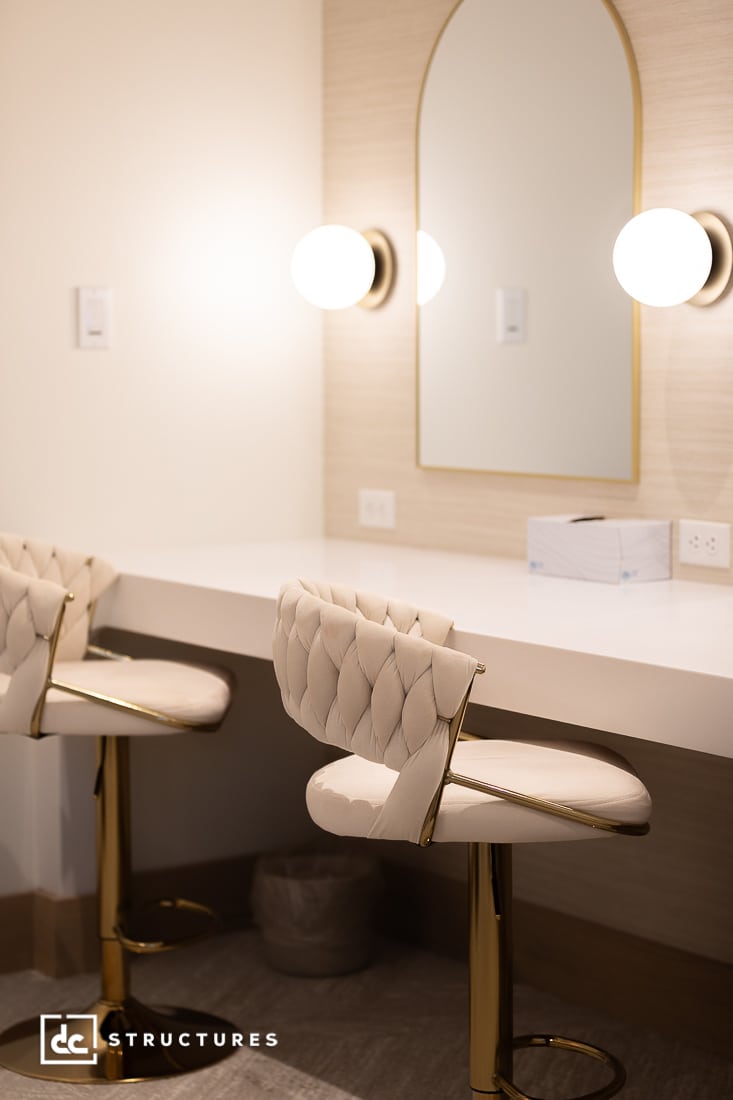 Modern vanity area with two tufted chairs with gold legs, white countertop, tall arched mirror, round wall lights; tissue box and trash bin are visible.