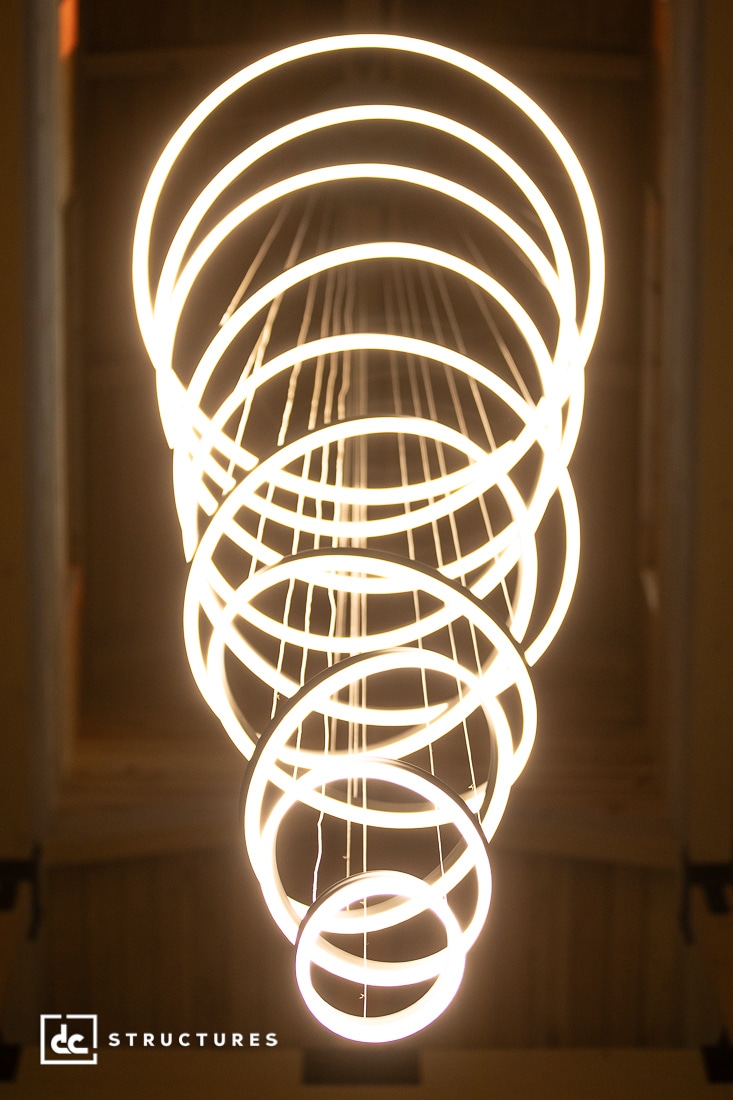 A modern chandelier of glowing circular rings of light spirals vertically; its bright glow stands out against a dim background.