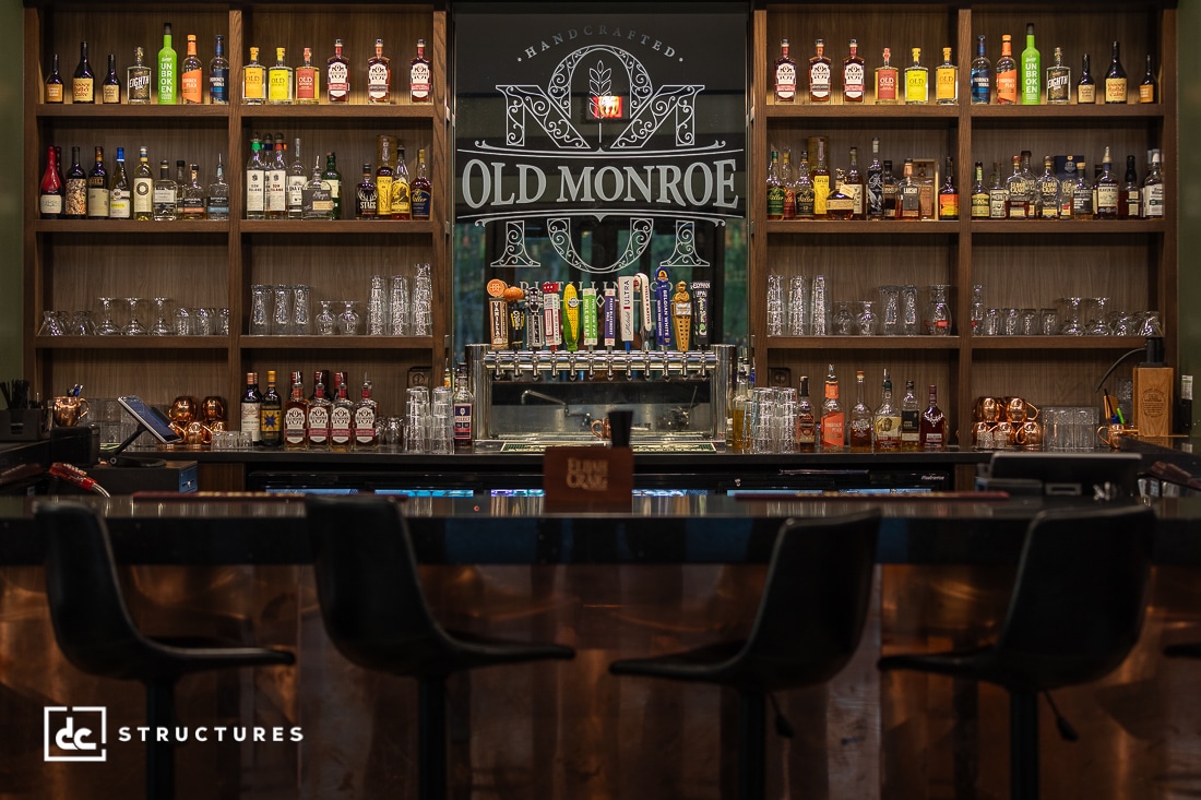 A modern bar with shelves of assorted liquor bottles and taps for draft beer. Four bar stools line the polished counter in the foreground.