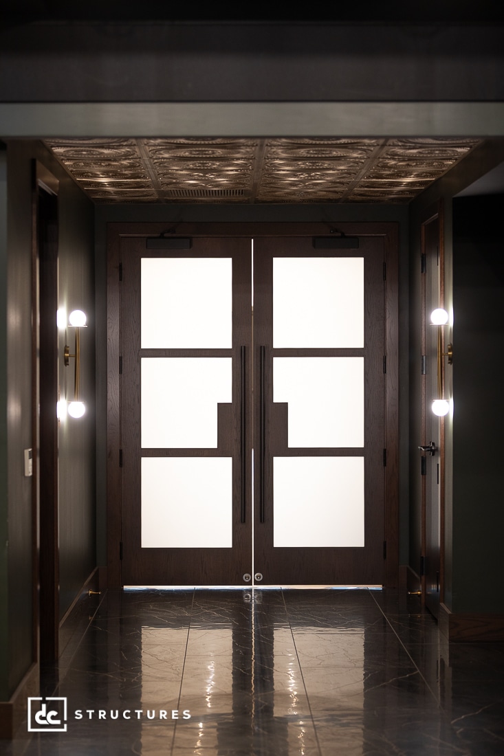 Double wooden doors with geometric frosted glass panels are centered in a hallway with dark walls, glossy tile floors, modern wall sconces, and a patterned ceiling.