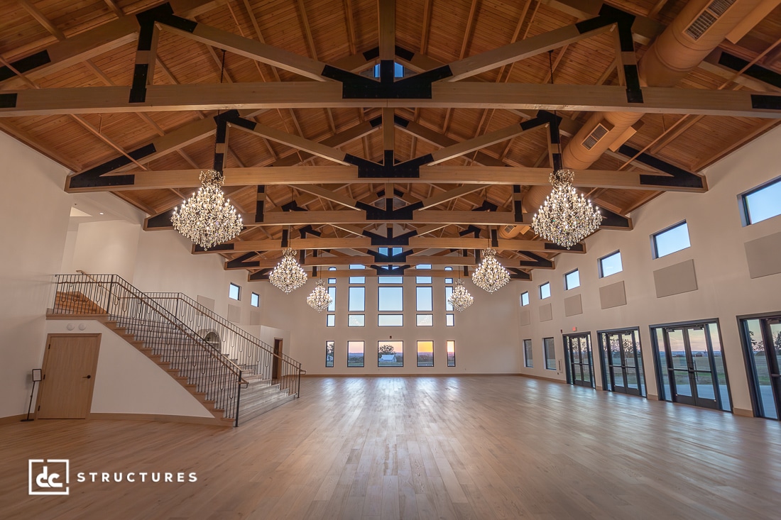A spacious, elegant hall with exposed wooden beams, multiple chandeliers, large windows, light wood flooring, a staircase left.