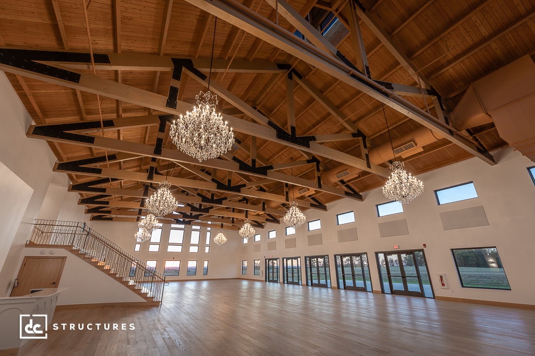 Spacious event hall with high wooden beams, chandeliers, large windows, and a staircase. The room has wood flooring and natural light streaming through the windows.