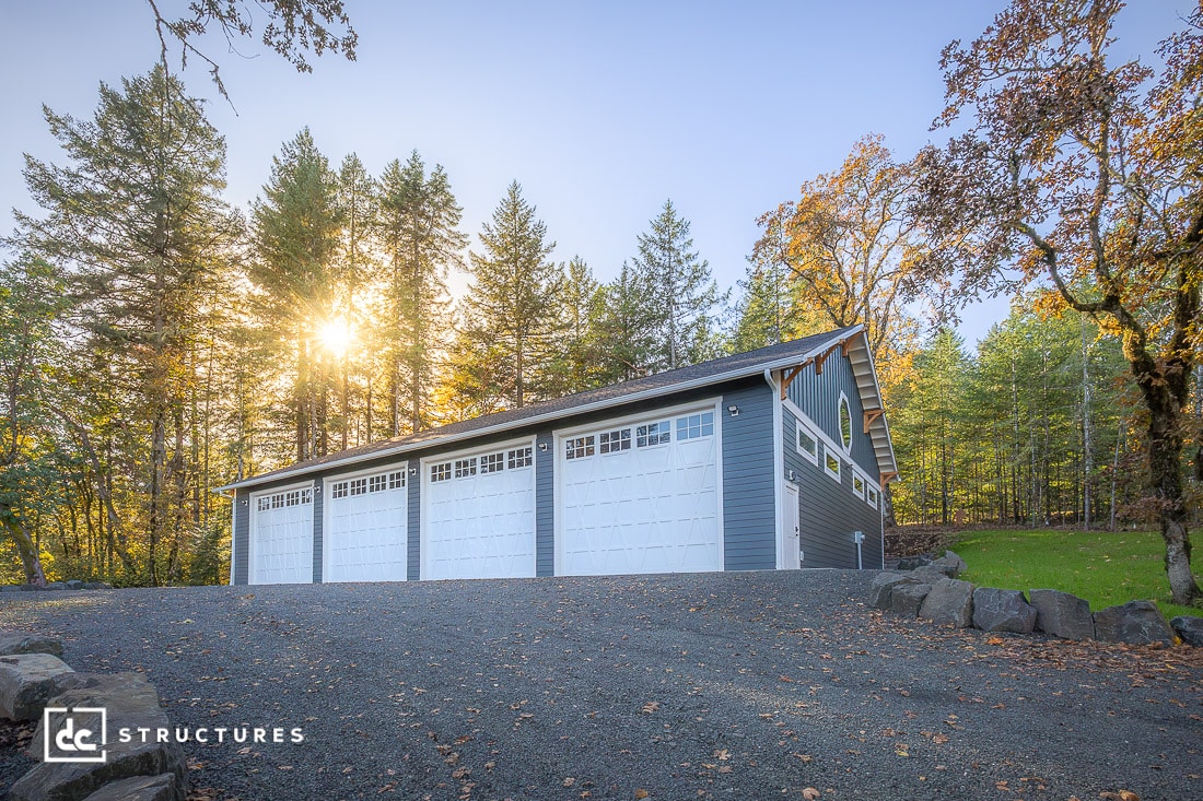 A large, gray four-car garage with white doors sits on a gravel driveway, surrounded by tall trees and green grass, with sunlight streaming through the forest in the background.