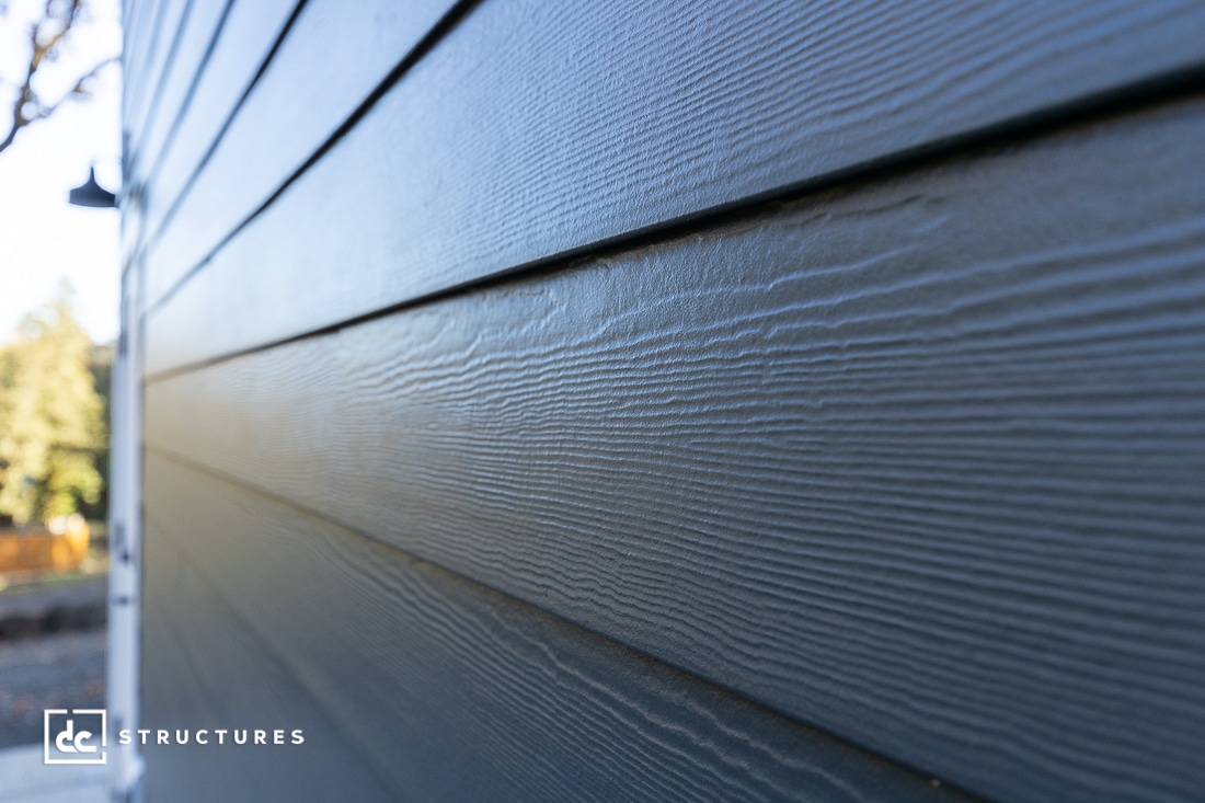 Close-up view of gray wood-textured siding panels on a building exterior, with visible grain patterns and sunlight casting subtle shadows. A blurred background includes trees and part of a lamp post.