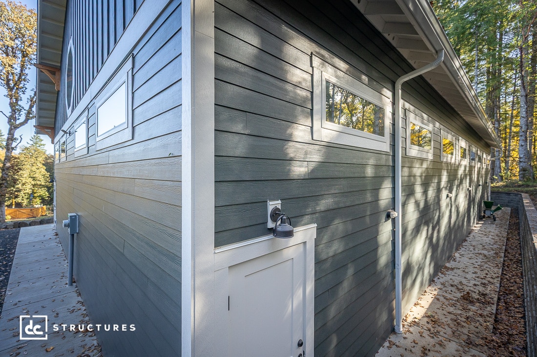 A modern green building with horizontal siding, tall narrow windows, a small white door, and exterior lighting, surrounded by trees and a concrete walkway.