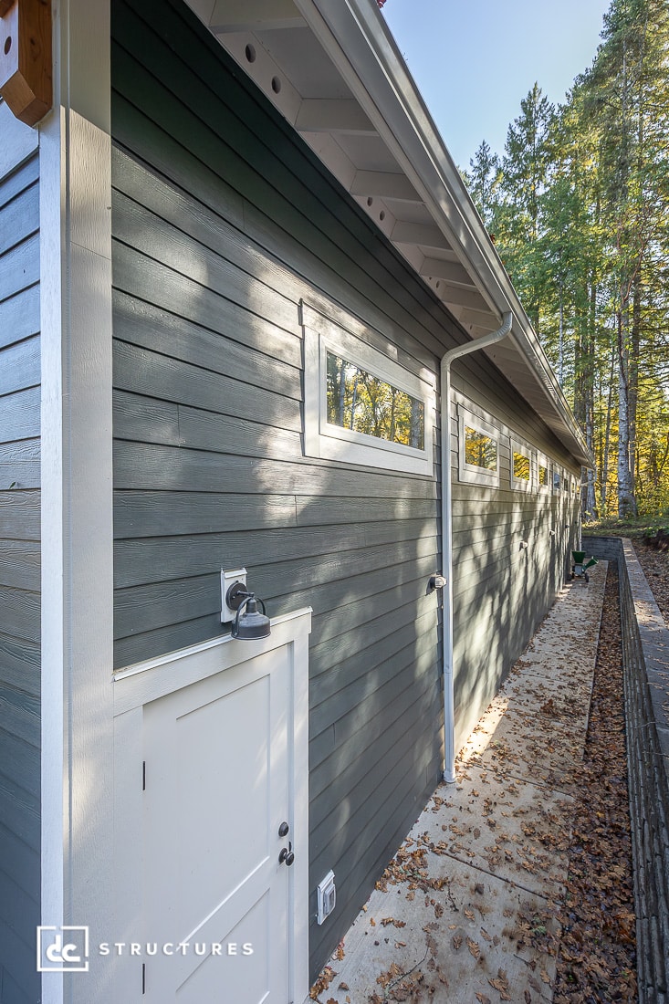 A long modern gray building with horizontal siding, narrow windows, a white door and trim, next to a forest with fallen leaves.