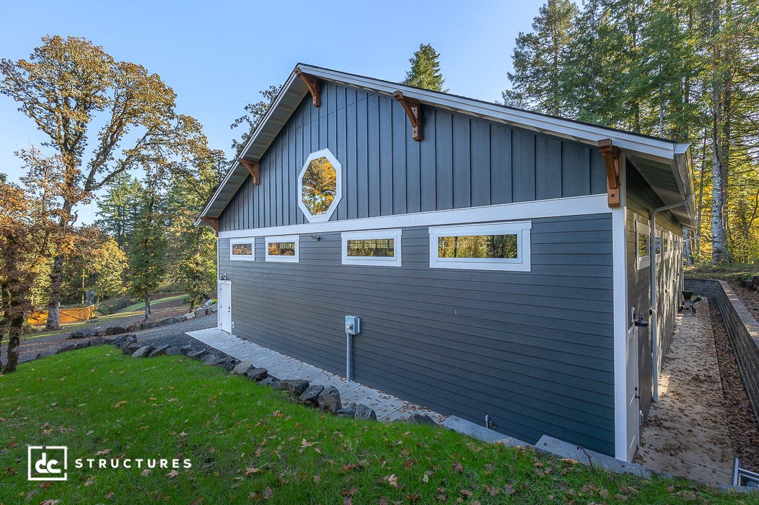 A modern gray and white barn-style building with large garage doors, surrounded by trees and greenery, under a clear blue sky.
