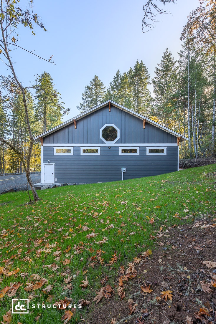 A gray barn-style building with white trim and a large octagonal window, on a grassy lawn with autumn leaves and trees under a clear sky.