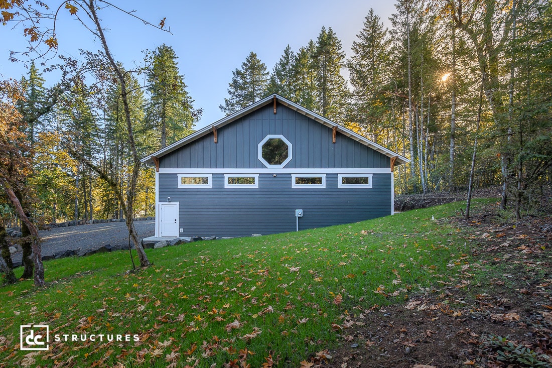 A modern gray barn with a sloped roof, white trim, and multiple windows stands on a grassy lawn surrounded by tall trees. Sunlight filters through the foliage.