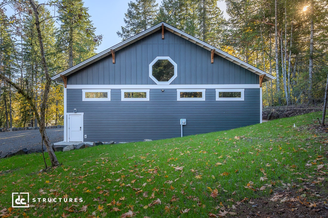 A gray barn-style building with white trim sits on a grassy hill among tall trees, featuring rectangular and octagonal windows.