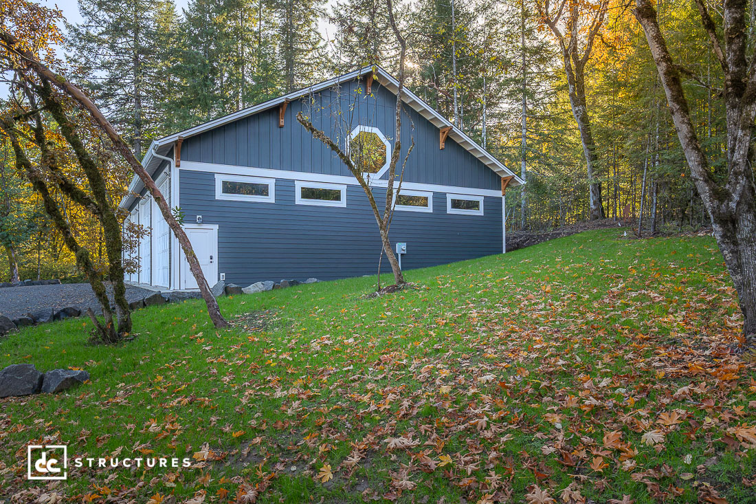 A blue and white barn-style building with a circular window sits on a grassy slope among trees with fallen autumn leaves. Sunlight filters through the trees.