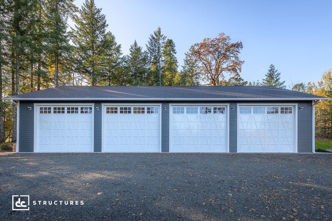 A large gray garage building with four white doors, each with geometric window designs, surrounded by trees on a gravel driveway.