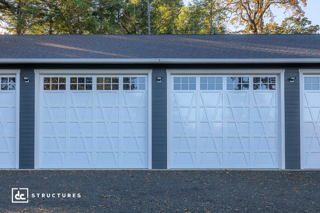 A modern gray and white garage building with three large white paneled doors featuring geometric patterns and windows, surrounded by trees.