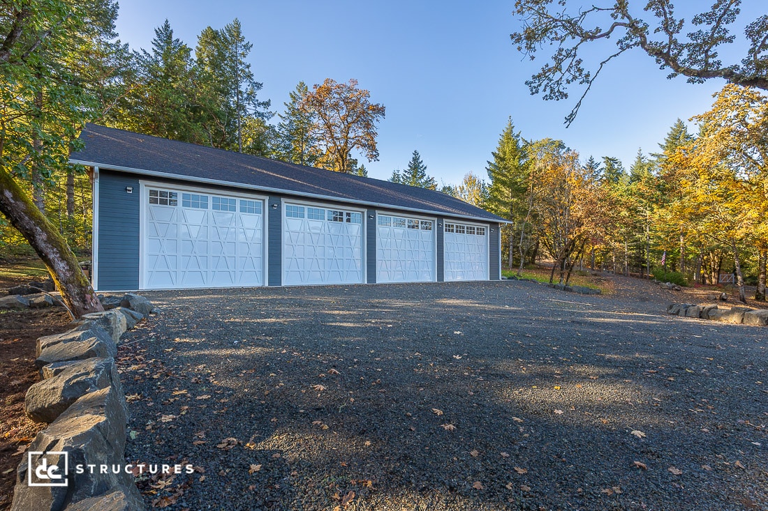 A large blue four-car garage with white doors sits on a gravel driveway, surrounded by trees and greenery on a clear, sunny day.
