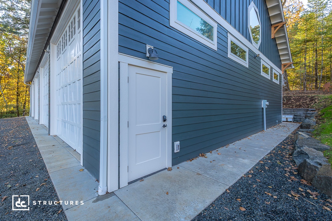 Side view of a modern blue building with white trim, white door, multiple windows, paved walkway, gravel area, and autumn trees.