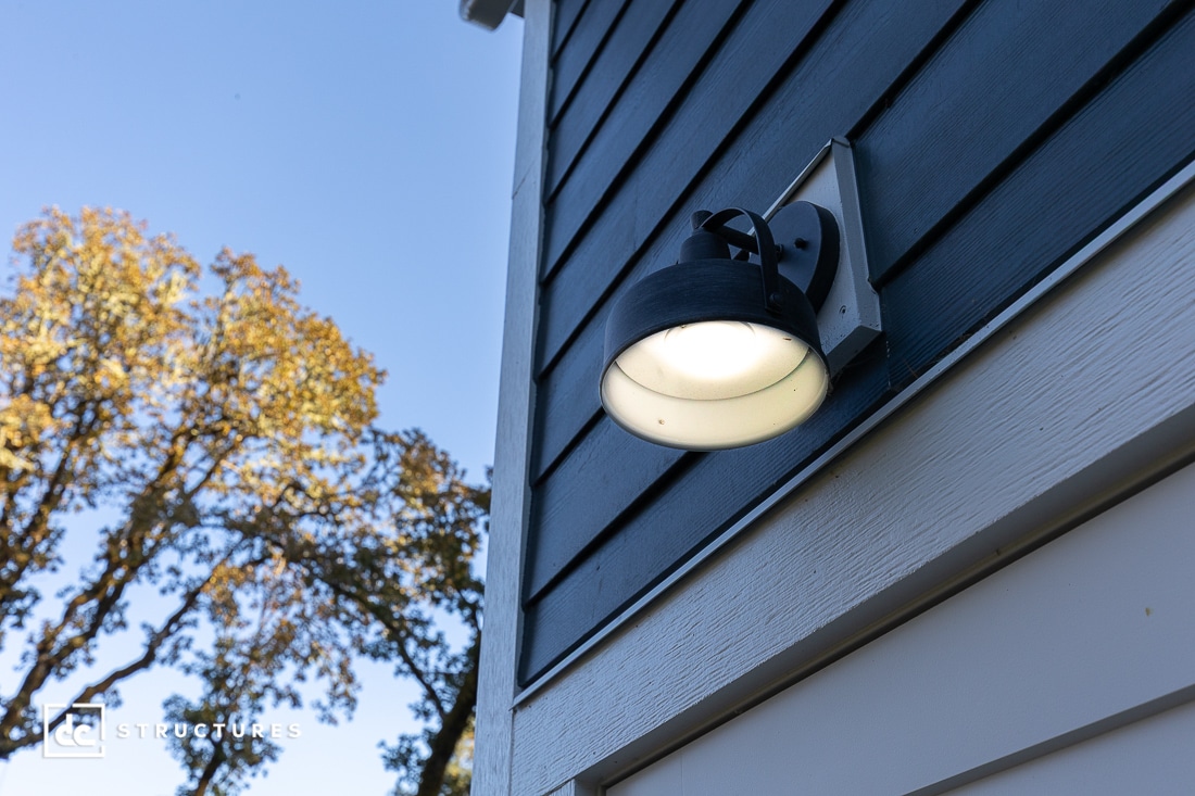 A close-up of a modern black outdoor wall light mounted on the dark blue siding of a house, with trees and a clear sky in the background.