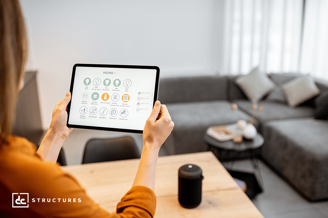 A person holds a tablet displaying a home automation interface while sitting at a table in a modern living room. A smart speaker and sofa are visible in the background.