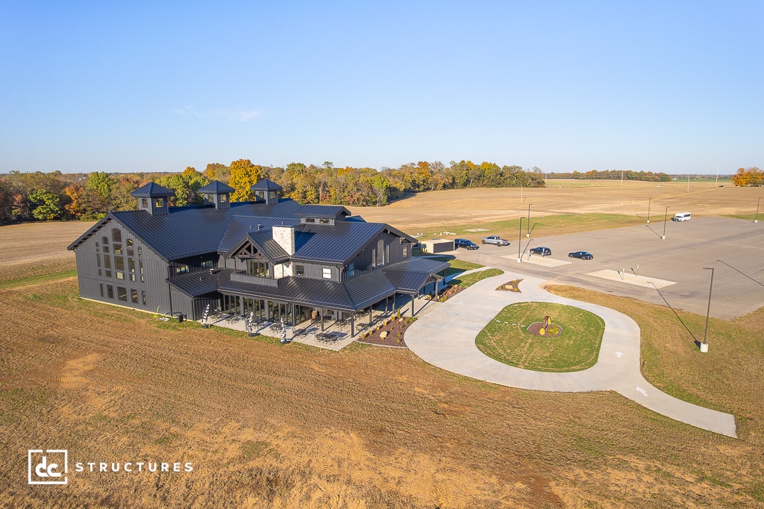 A large, modern barn-style building with a black metal roof sits by a curved driveway and parking lot, surrounded by fields and trees. Several cars are parked nearby under a clear sky.