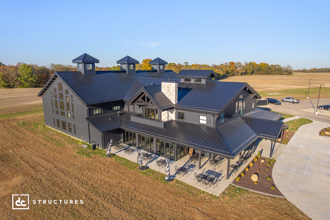 A large modern barn-style building with black metal roofing, cupolas, open fields, outdoor seating, and a mix of wood and stone.