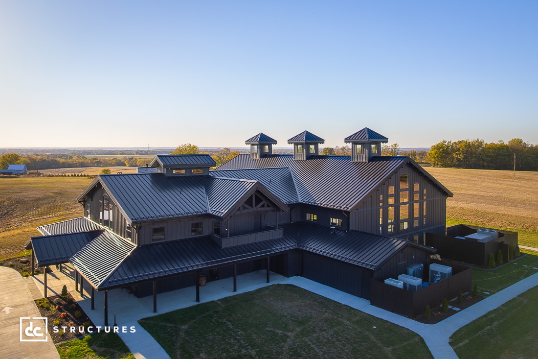 A large modern barn-style building with a dark metal roof, multiple cupolas, and large windows in a rural landscape under a blue sky.