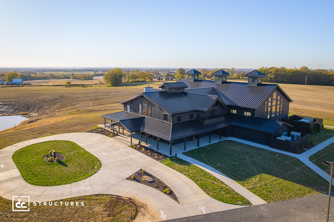 A large, modern barn-style building with dark metal roofing sits on a spacious property with driveways, surrounded by open fields.