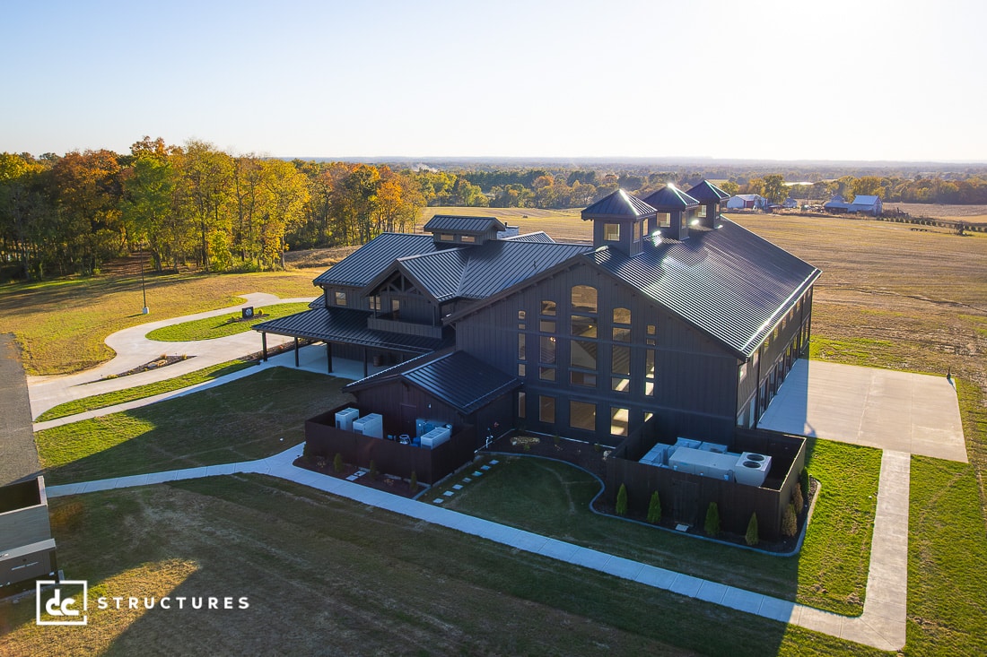 A large modern barn-style building with black siding and cupolas sits on a grassy property surrounded by trees and open fields.