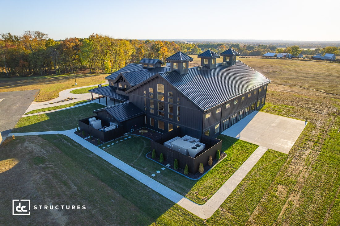 A large modern black barn with multiple cupolas, big windows, and a metal roof sits on an open lot with grass, trees, and a driveway.