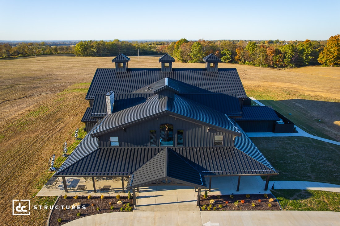 Aerial view of a large, modern barn-style building with a black metal roof and three cupolas, surrounded by fields and trees under a clear sky.