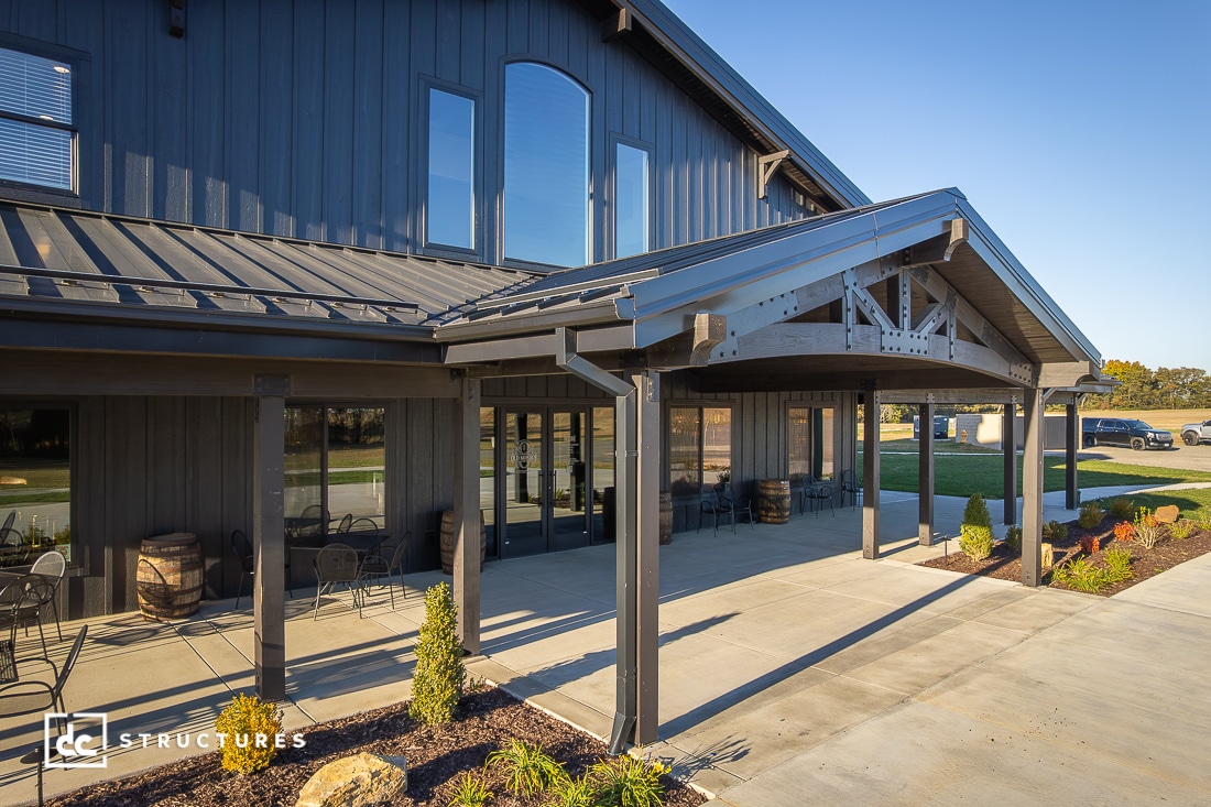 A modern barn-style building with a metal roof, large glass doors, covered entryway, outdoor seating, plants, and sunny sky.