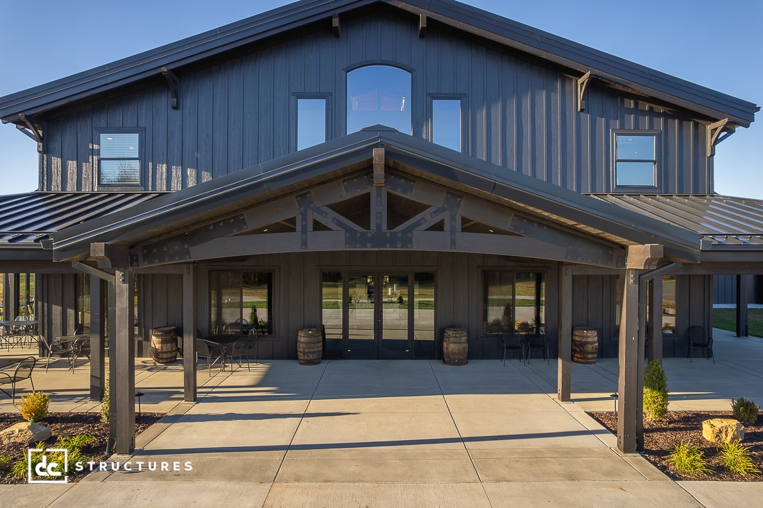 A modern black barn-style building with large windows, a covered entrance supported by wooden beams, patio seating, double doors, and two barrels flanking the entry.