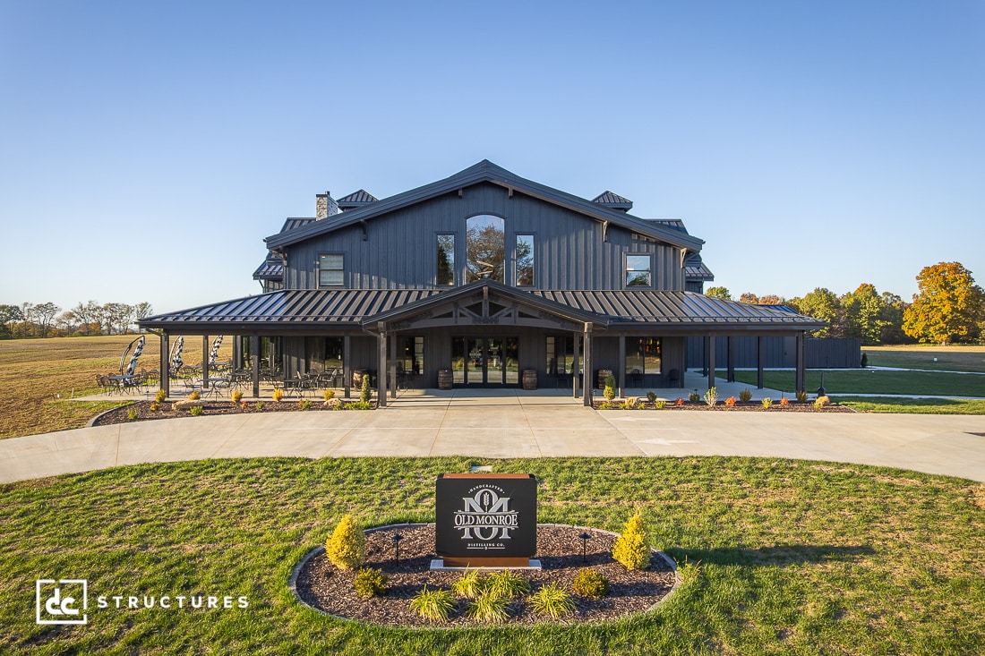 A large, modern barn-style building with dark siding and a wide covered entrance, surrounded by landscaped grass and a circular driveway. Trees and open fields are in the background.
