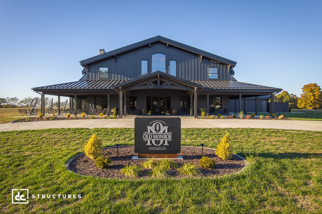 A large modern barn-style event venue with dark siding and a covered entrance sits under a clear blue sky. The surrounding lawn is landscaped with shrubs.