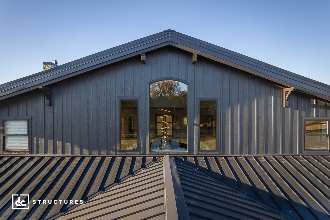 A close-up of a modern dark gray barn-style building with vertical wood siding, large central window, and metal roof, set against a clear blue sky.