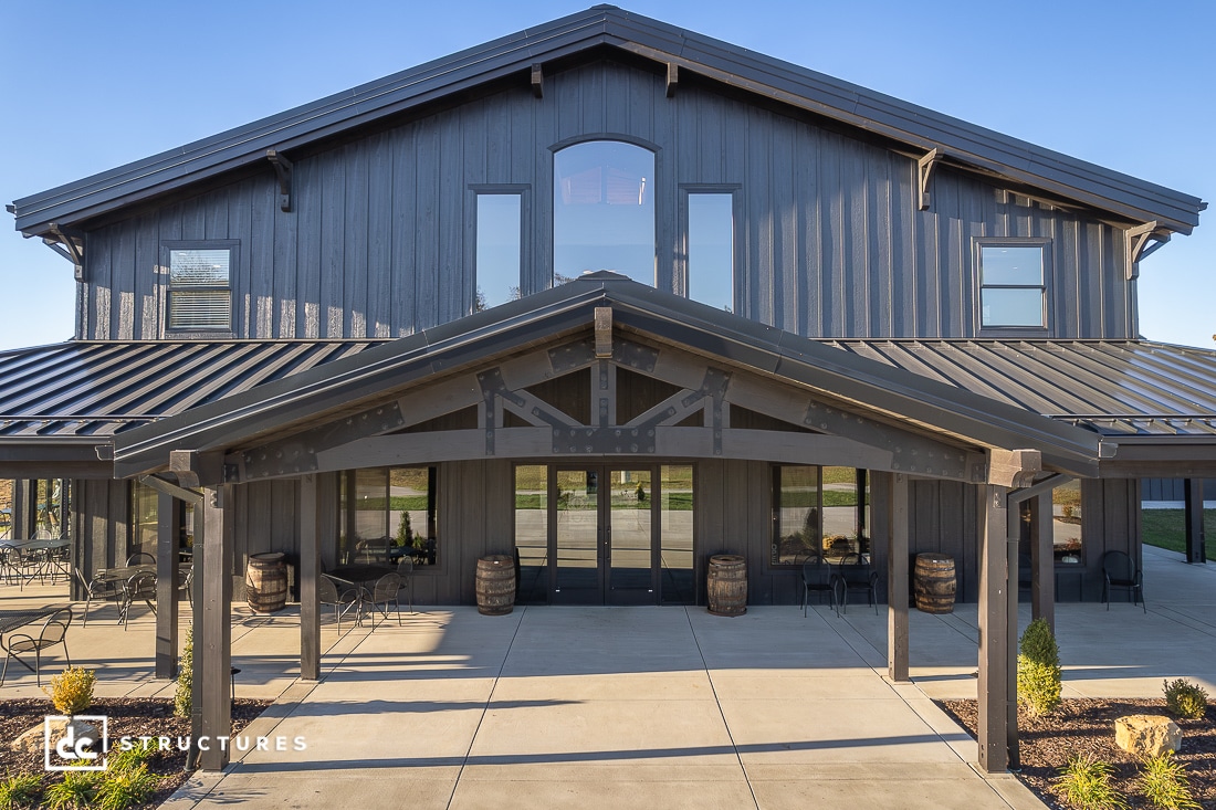 A modern dark gray barn-style building with large windows, a metal roof, and a covered entrance. Outdoor chairs and barrels in front.
