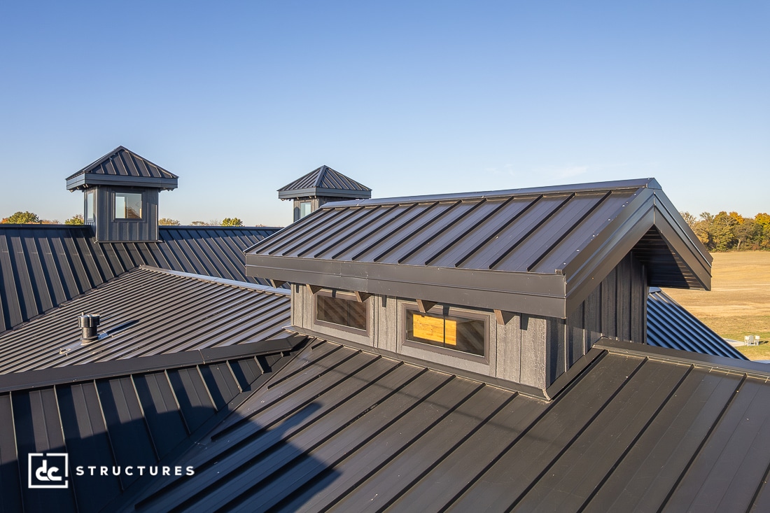 A close-up view of a modern black metal roof with dormer windows and cupolas, set against a clear blue sky and open field.