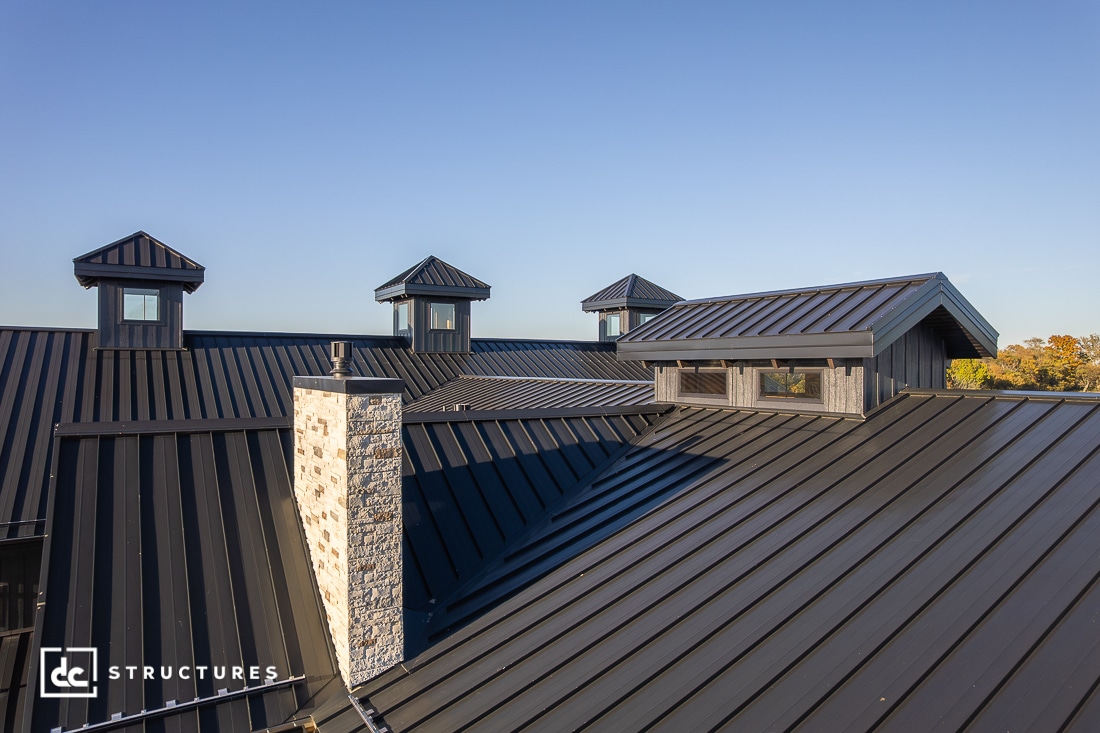 Modern building with a black metal roof, stone chimney, and several cupolas against a clear blue sky.