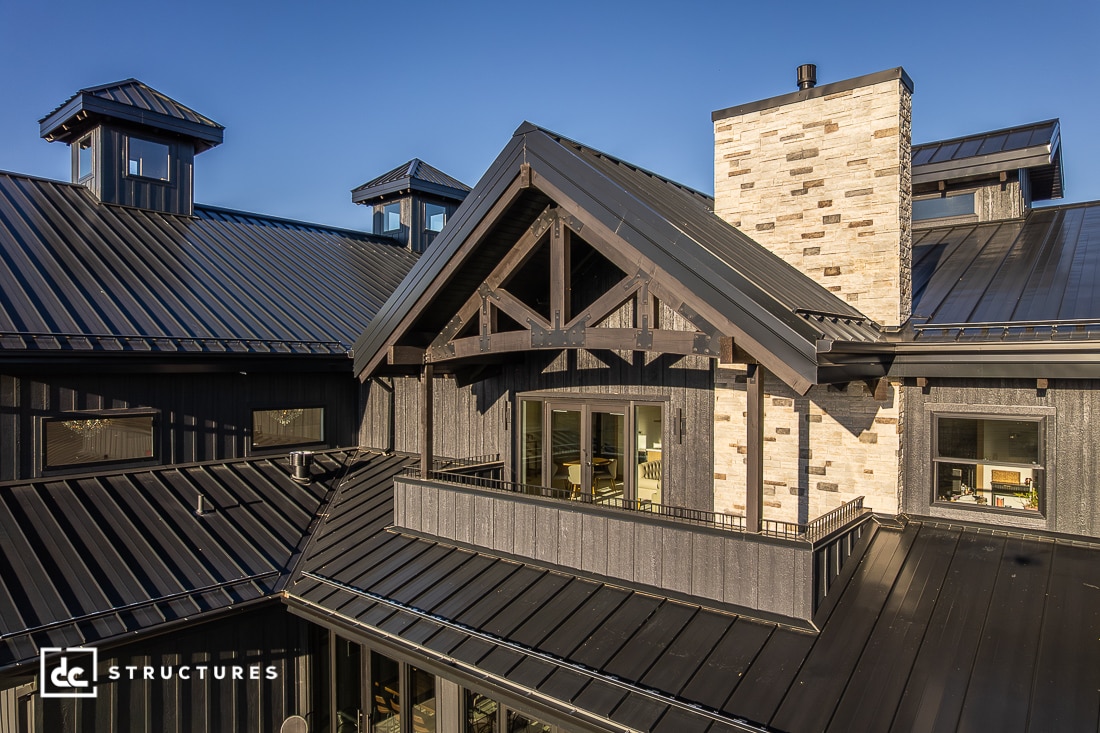 Modern black metal-roofed home with exposed timber trusses, stone chimney, and large windows, viewed in sunlight.